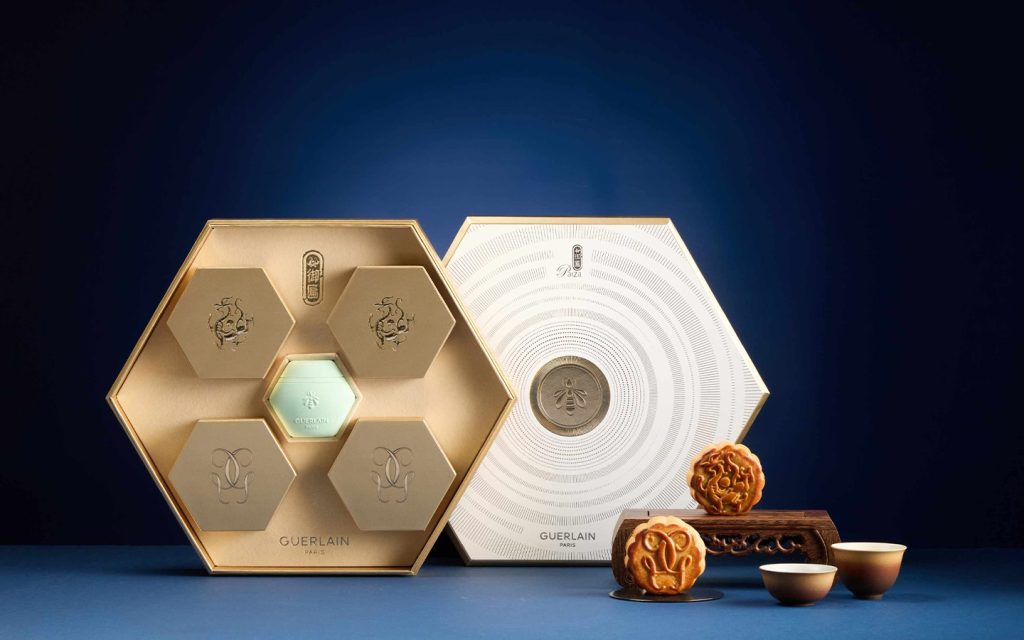 Four honey-infused mooncakes arranged around a Guerlain hand cream in a honeycomb tray, the bee emblem echoed on the lid